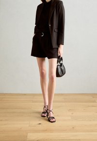 Blazer noir ajusté sur un top noir, associé à un short noir. Des sandales noires à lanières et un petit sac noir complètent la tenue.