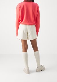 Felpa a maniche lunghe color corallo, pantaloni corti color crema, calze sopra il ginocchio color crema, scarpe beige. Abbigliamento casual con vestibilità rilassata e morbida texture.