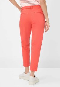 Pantaloni corallo con taglio slim e lunghezza alla caviglia, dotati di tasche posteriori. Indossati con sneakers chiare e una maglietta a righe.