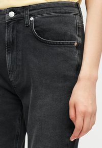 Κοντινό πλάνο ενός ατόμου που φορά μαύρο τζιν παντελόνι με ορατή μπροστινή τσέπη, κουμπί και ένα χέρι που στηρίζεται δίπλα στο πόδι.