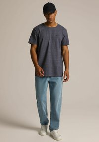 Szara koszulka z krótkim rękawem z kieszonką na piersi, w komplecie z jasnoniebieskimi dżinsami i białymi sneakersami, noszona z czapką w kolorze czarnym.