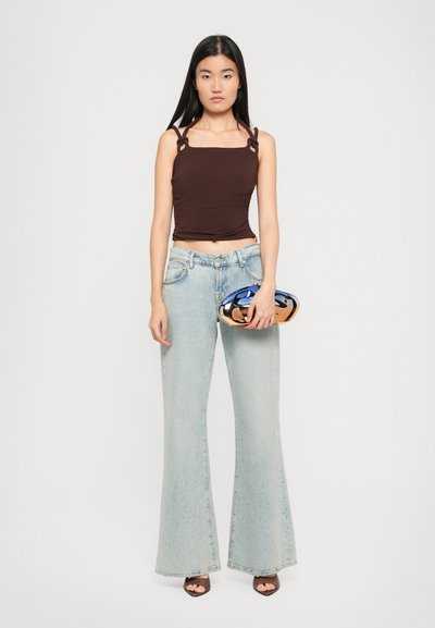 Bruine cropped top met vierkante halslijn en gedraaide bandjes, gecombineerd met lichtblauwe flaired jeans. Model houdt een glanzende blauwe en gouden handtas vast.