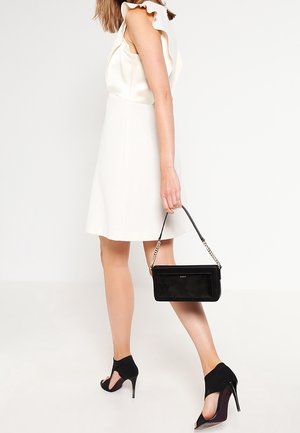 Femme en robe blanche sans manches et talons hauts noirs tenant un sac à main noir avec une bandoulière chaîne, marchant devant un fond uni.