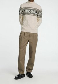 Man med beige kabelstickad tröja med mönstrad bröststripe, bruna veckade byxor och svarta läderskor på vit bakgrund.