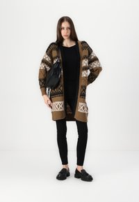 Larga chaqueta de punto en tonos marrones, negros y blancos con patrones geométricos, combinada con una blusa negra, pantalones ajustados negros y zapatos oscuros.