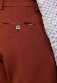 Roodbruin gekleurde broek van een gestructureerde stof, met een knoopsluiting en een achterzak. Een deel van een geruit overhemd is zichtbaar boven de broek.