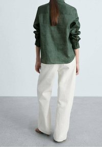 Camicia di lino verde con una vestibilità rilassata, maniche arrotolate e yoke posteriore. Abbinata a pantaloni ivory a gamba larga, che mostrano un look informale.