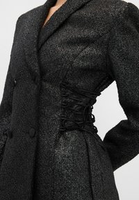Blazer nero con texture, dal design doppiopetto, caratterizzato da spalle a sbuffo e un laccio in vita per una vestibilità regolabile.