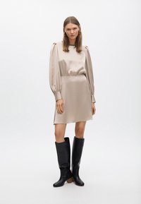 Vestido satinado beige con mangas abullonadas y cintura ajustada, combinado con botas de cuero negras de caña alta y tacón corto.
