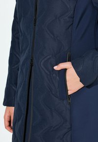 Person som bär en mörkblåtexturerad jacka med hand i det dragkedjeförsedda sidofacket, som visar detaljer om frontdragkedjan och ärmarna.