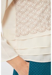 Blusa de crema con bordado geom�étrico en un patrón floral repetido, con paneles horizontales superpuestos y una textura suave. Tela teal visible por debajo.