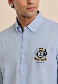 Camisa de botones con rayas azules y blancas, que presenta un logo náutico bordado en el pecho. Tejido de algodón, cuello estándar y cierre de botones.