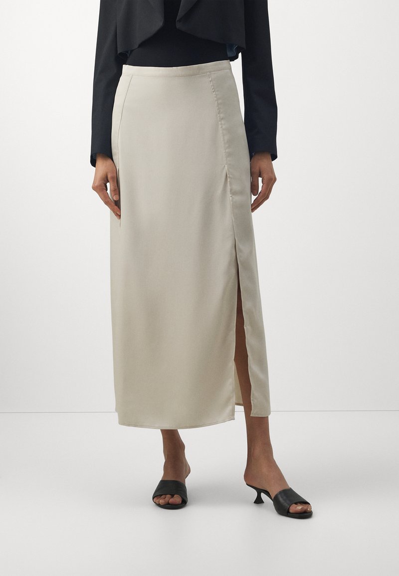 Gonna maxi in satin beige chiaro con uno spacco laterale, vita alta e texture morbida, abbinata a mule nere con punta aperta.