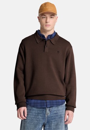 Jonge man die een bruine trui met kraag draagt over een blauw geruit overhemd, lichtblauwe spijkerbroek en een beige Timberland pet, staand tegen een effen achtergrond.