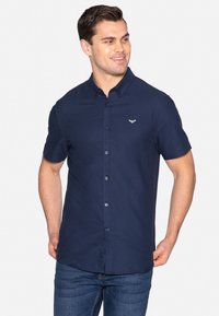 Marineblaues kurzarm Hemd mit Knopf und Kragen, mit einem Logo auf der Brust und blauen Knöpfen, getragen mit blauen Jeans.