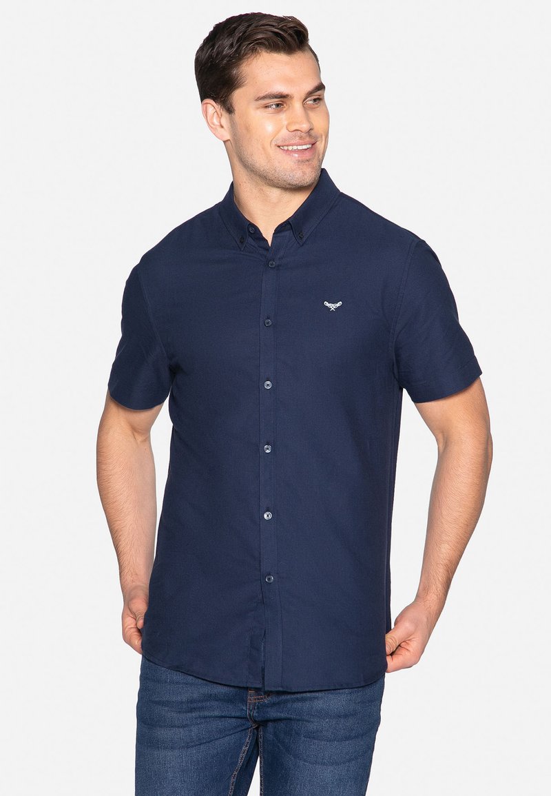 Marineblaues kurzarm Hemd mit Knopf und Kragen, mit einem Logo auf der Brust und blauen Knöpfen, getragen mit blauen Jeans.