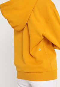 Żółta bluza z kapturem noszona przez osobę w białych spodniach, pokazująca widok z tyłu i z boku z małym białym logo po boku.