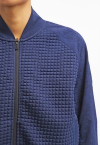 Chaqueta azul marino con cremallera, con un patrón acolchado texturizado en el frente, mangas suaves y cuello alto con un detalle de cremallera negra.