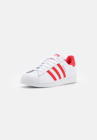 Zapatilla blanca con acentos rojos. Presenta tres rayas rojas, una puntera de goma texturizada y una suela blanca sólida. Diseño con cordones.