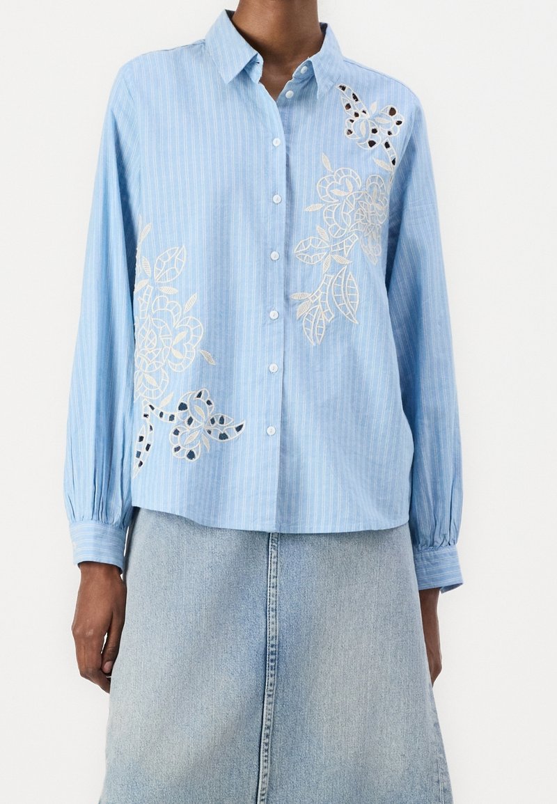 Chemise à rayures bleu clair boutonnée avec broderie florale blanche portée avec une jupe en denim clair, montrée du cou jusqu'à mi-cuisse.