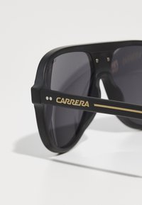 Ochelari de soare negri cu un finisaj mat, lentile întunecate și un logo aurit "CARRERA" pe ramă. Prezintă un design unghiular distinctiv.