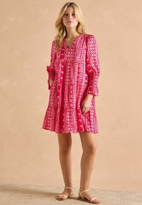 Różowa i biała sukienka w wzory z dekoltem w serek, długimi rękawami i falbanowym dołem; wykonana z lekkiego materiału o luźnym kroju.