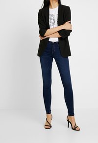 Svart blazer över en vit t-shirt med tryck, kombinerad med mörkblå skinny jeans och svarta högklackade sandaler med remmar. Smal passform och avslappnad design.