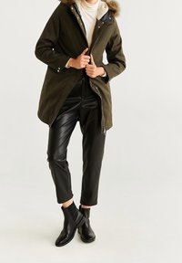 Oliwkowa parka z kapturem wyłożonym futerkiem, w zestawieniu z czarnymi spodniami z imitacji skóry i czarnymi botkami na kostkę. Płaszcz posiada zamek błyskawiczny oraz zatrzaski.