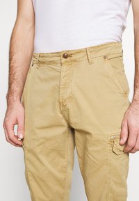 Beige cargo hlače iz mehkega bombaža, s sprednjim gumbom za zapiranje, stranskimi žepe in stranskim žepom z zavihkom ter šivanjem.