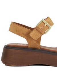 Tan suède sandaal met een verstelbare gesp en een platformzool, met een gladde textuur en een contrasterende bruin houtachtige basis.
