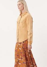 Lichtgele denim blouse met knopen en twee borstzakken, gecombineerd met een gemusterde bruine rok met kleurrijke vormen.