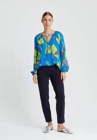 Blouse bleue avec des motifs abstraits verts et roses, manches longues et un col à cordon, associée à un pantalon noir fuselé et des ballerines métalliques.