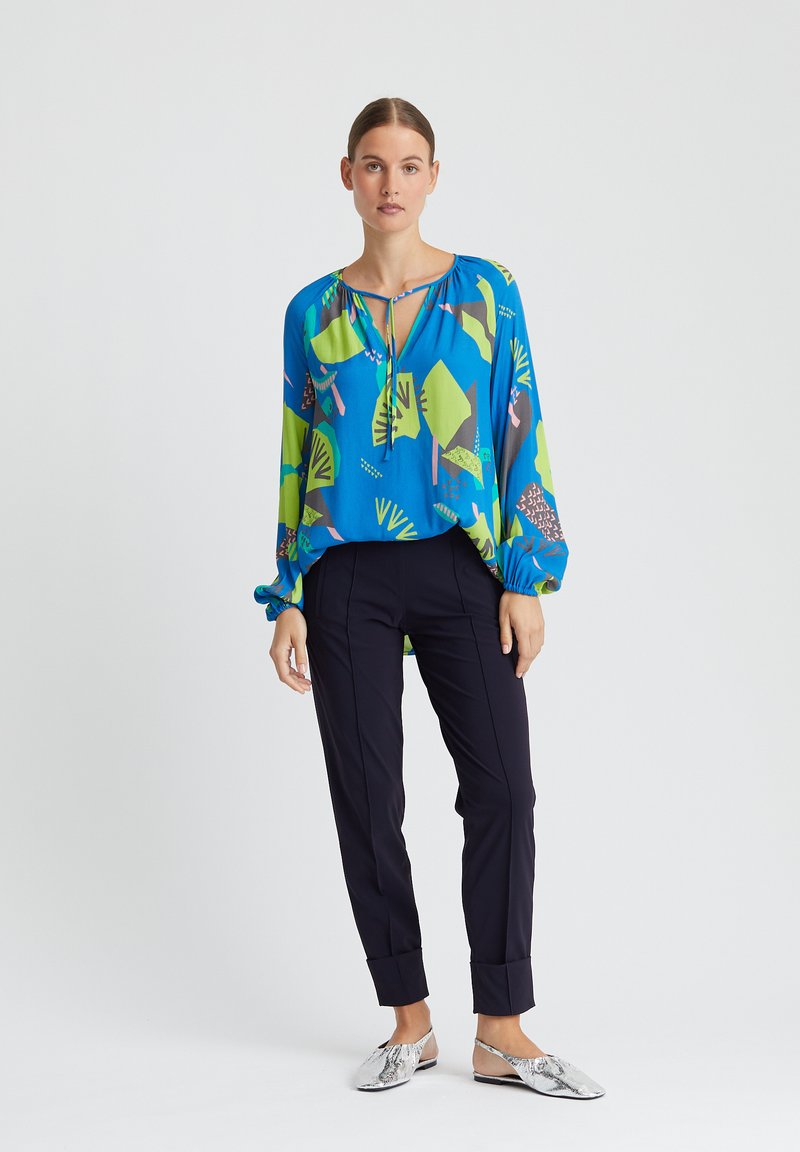 Blouse bleue avec des motifs abstraits verts et roses, manches longues et un col à cordon, associée à un pantalon noir fuselé et des ballerines métalliques.