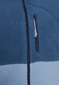 Modrá flísová bunda s tmavě námořnickým vertikálním zipem a zipovou náprsní kapsou, která má bílý a modrý stahovací provázek.