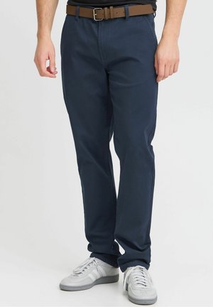 Mężczyzna ubrany w granatowe spodnie, białe trampki, brązowy pasek i czarną koszulę na jednolitym białym tle.