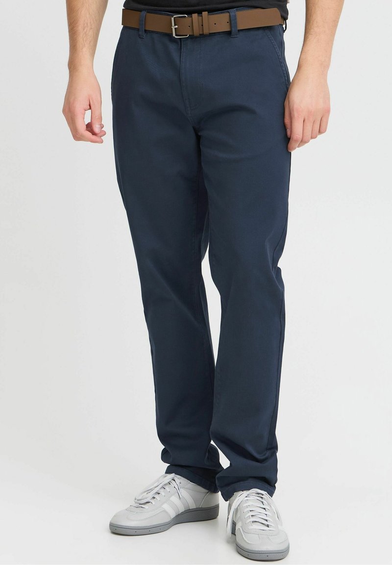 Homme portant un pantalon bleu marine, des baskets blanches, une ceinture marron et une chemise noire sur un fond blanc uni.