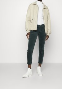 Beige bomberjakke med lynlås, hvid turtleneck, mørkegrøn kordfløjl-bukser og hvide højhælede sneakers. Glat stof, tætsiddende pasform.