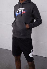 Šedá mikina s kapucí a přední kapsou, na které se nachází vícolorový logo Nike. Černé kraťasy s velkým bílým logem Nike na boku.