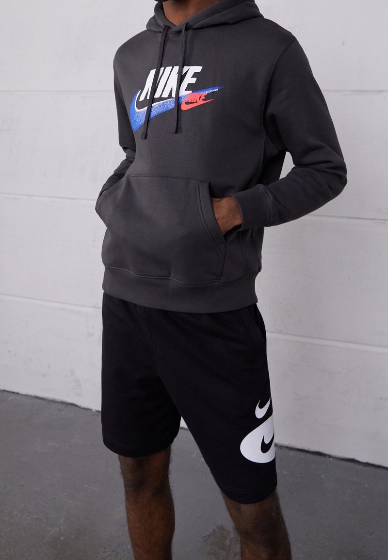 Šedá mikina s kapucí a přední kapsou, na které se nachází vícolorový logo Nike. Černé kraťasy s velkým bílým logem Nike na boku.