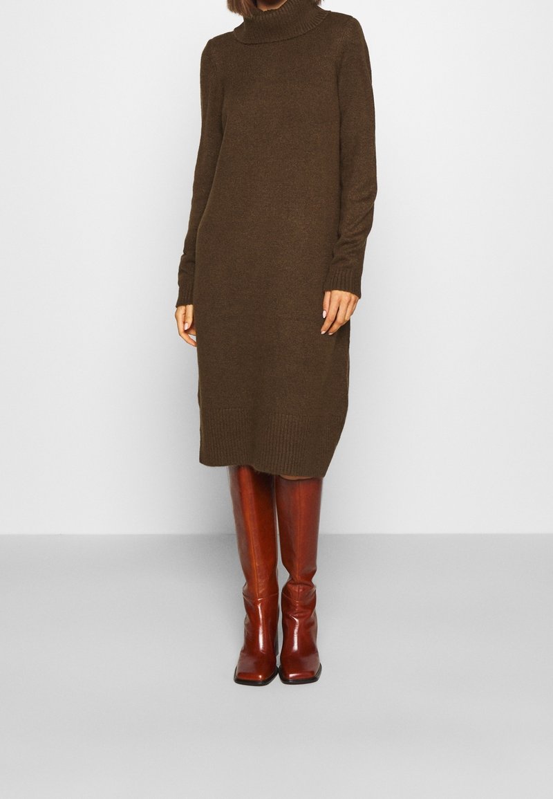 Robe en maille marron à col roulé avec manches longues, poignets et ourlet côtelés. Associée à des bottes hautes en cuir marron brillant.