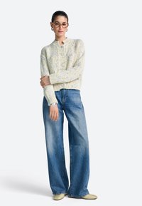 Gestrickte Strickjacke in pastellgelben und blauen Farbtönen, strukturiertes Design, kombiniert mit weiten blauen Jeans und pastellfarbenen Schuhen, ergänzt durch eine Brille.