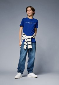 Modrá bavlněná tričko s logo "TOMMY HILFIGER", spárované s volnými džínami a béžovo-námorníkově pruhovaným svetrem uvázaným kolem pasu. Bílé tenisky.