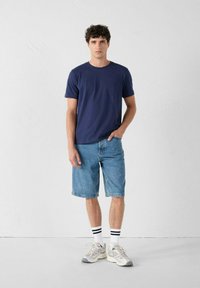 Navyblauwe katoenen t-shirt, relaxed fit, gecombineerd met lichtblauwe denim shorts. Witte sokken met zwarte strepen en grijze sportschoenen maken de look compleet.