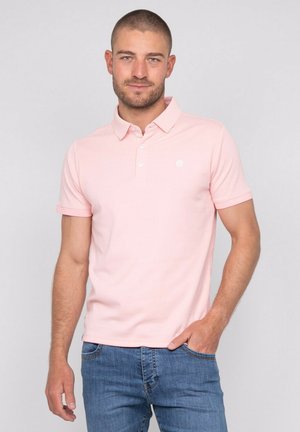 Homme aux cheveux courts et barbe portant un polo rose clair et un jean bleu, debout avec une main dans la poche devant un fond uni.