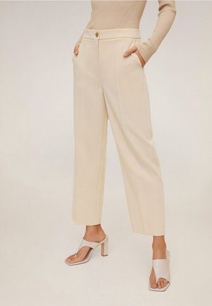 Femme portant un pantalon beige à jambes larges avec poches avant, associé à un haut beige côtelé à manches longues et des sandales beiges à talons ouvertes.