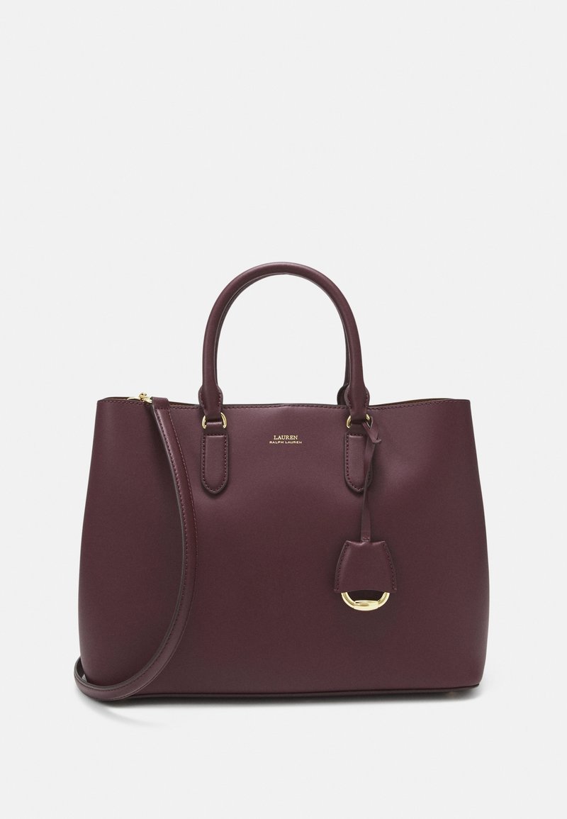 Sac à main en cuir violet foncé avec deux poignées, une longue bandoulière, et des accessoires dorés, avec une petite étiquette en cuir suspendue.