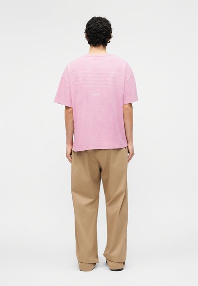 Άνδρας με σγουρά σκούρα μαλλιά, γυρισμένος με την πλάτη, φοράει ένα φαρδύ ροζ T-shirt με κείμενο στην πλάτη και φαρδιές μπεζ παντελόνες.
