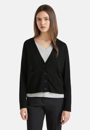 Zwarte cardigan met een V-hals, knopen aan de voorkant en twee borstzakken, gedragen over een grijze V-hals shirt, met lange mouwen en een getailleerd ontwerp.