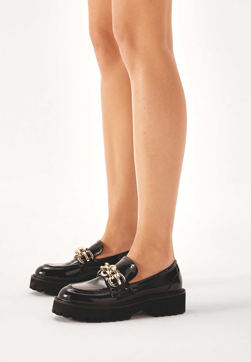 Black patent leather loafers with a chunky sole and gold chain detail on the front, featuring a smooth finish and rounded toe.
