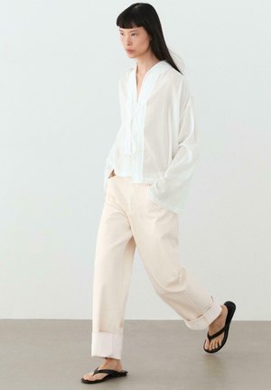 Femme marchant avec un chemisier blanc ample, un pantalon beige clair retroussé et des tongs noires sur un fond blanc uni.